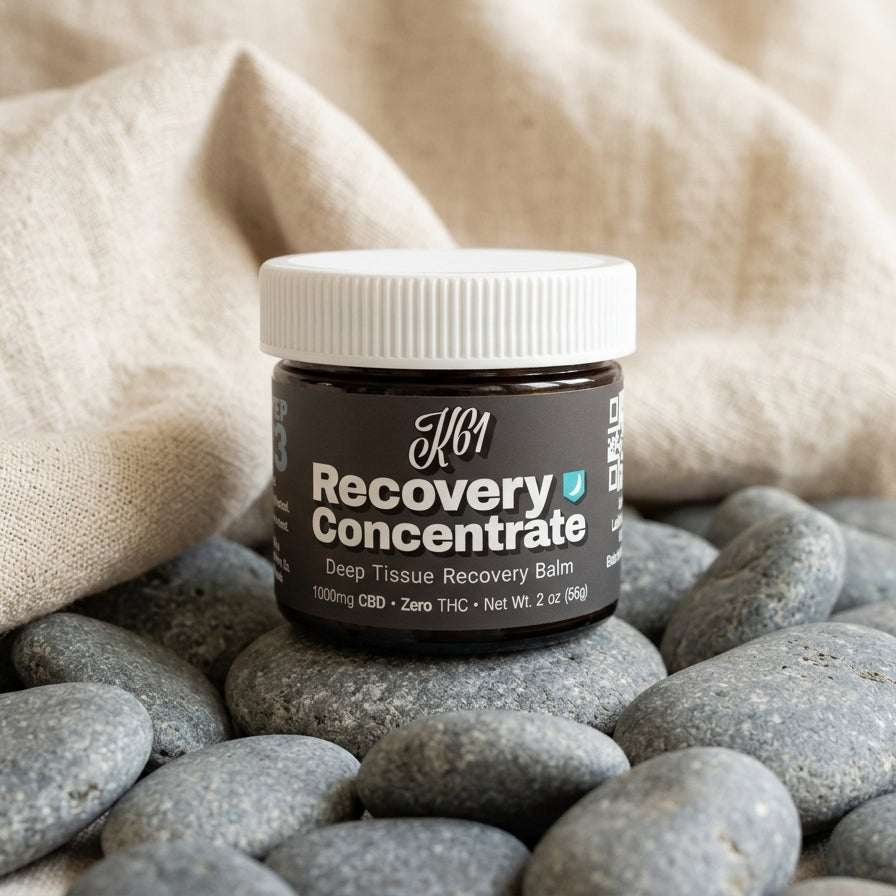 K61 Recovery Concentrate deep tissue balm jar resting on smooth river stones with natural linen