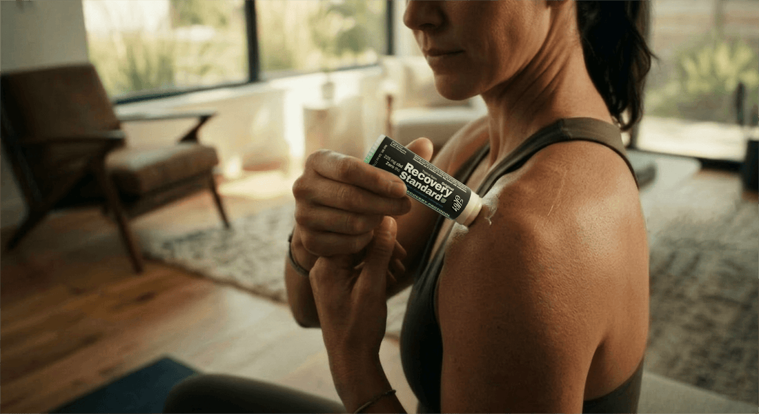A woman applying K61 Recovery Standard balm stick to her shoulder at home