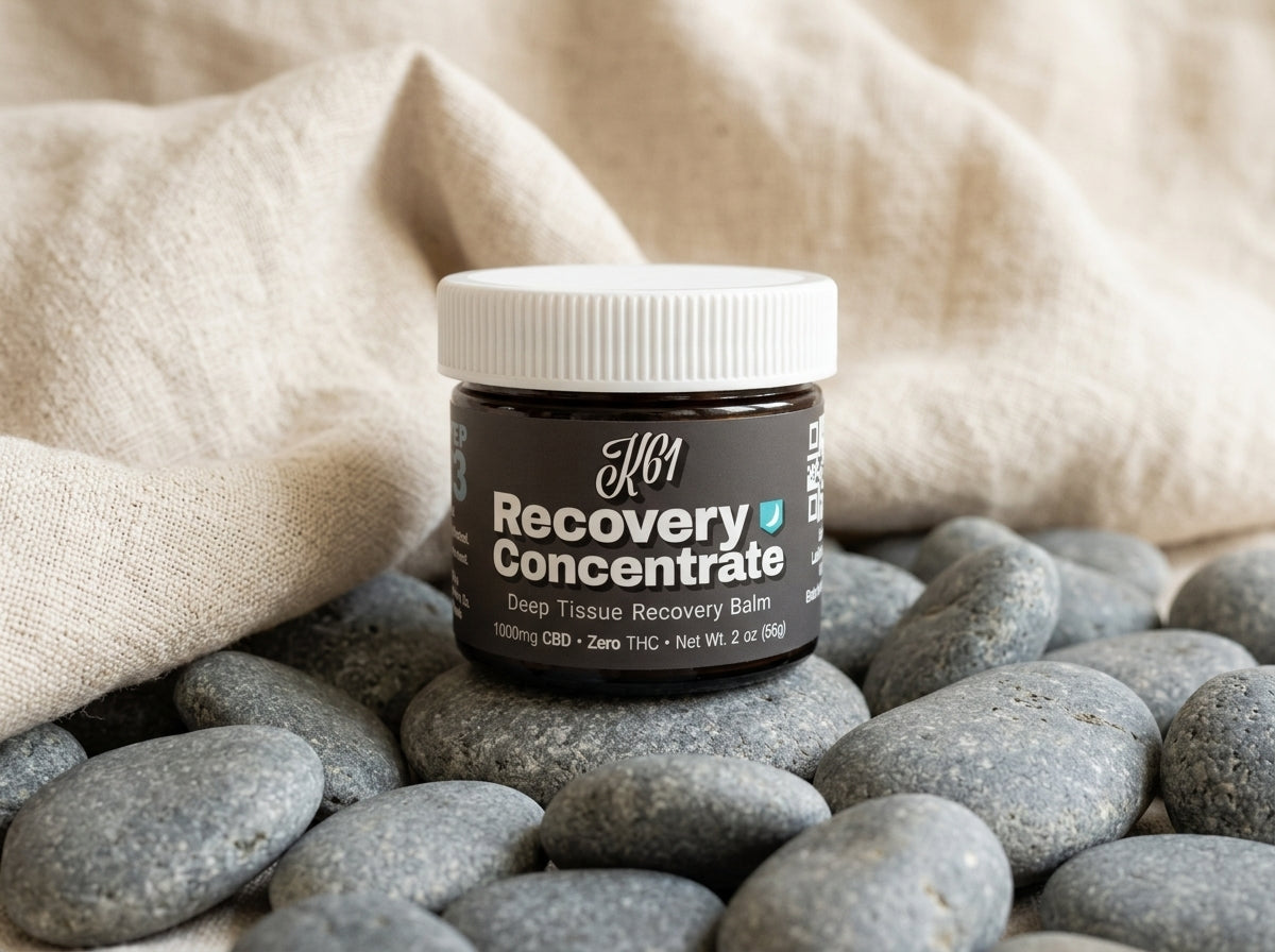 K61 Recovery Concentrate deep tissue balm jar resting on smooth river stones with natural linen