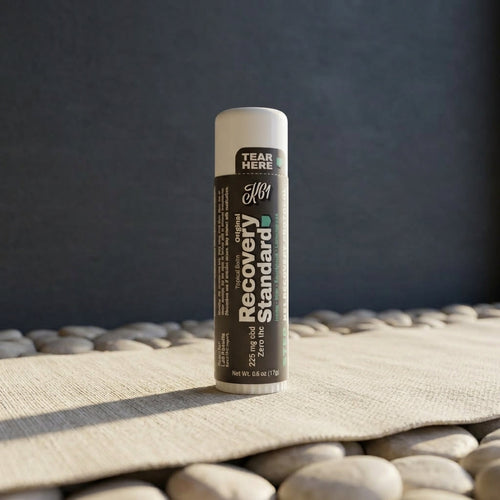 K61 Recovery Standard balm stick resting on smooth river stones with linen