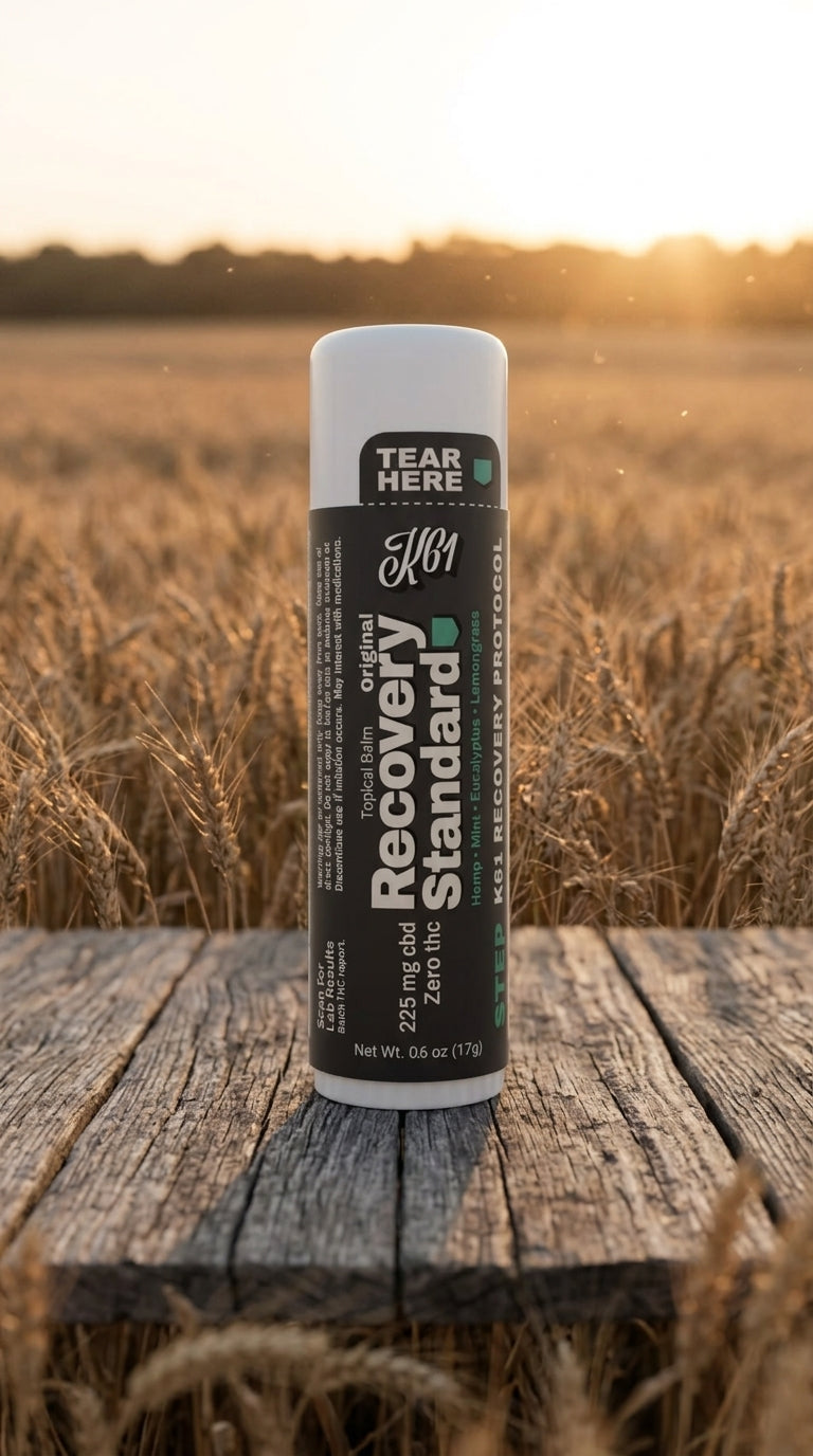 K61 Recovery Standard balm stick on weathered wood beside a Kansas wheat field at golden hour